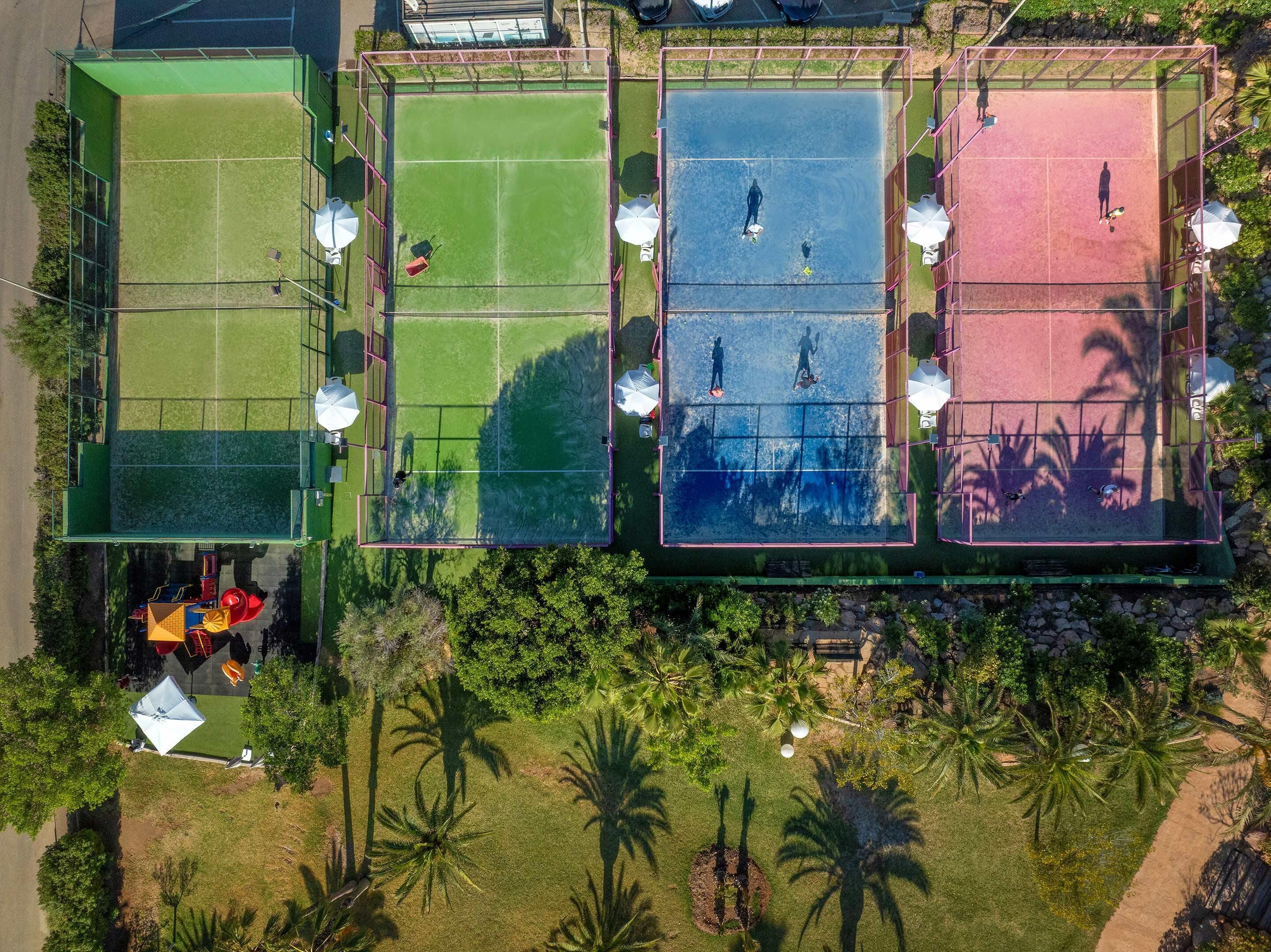 Luftaufnahme eines Sportkomplexes mit mehreren Tennisplätzen, umgeben von viel Grün und Palmen.