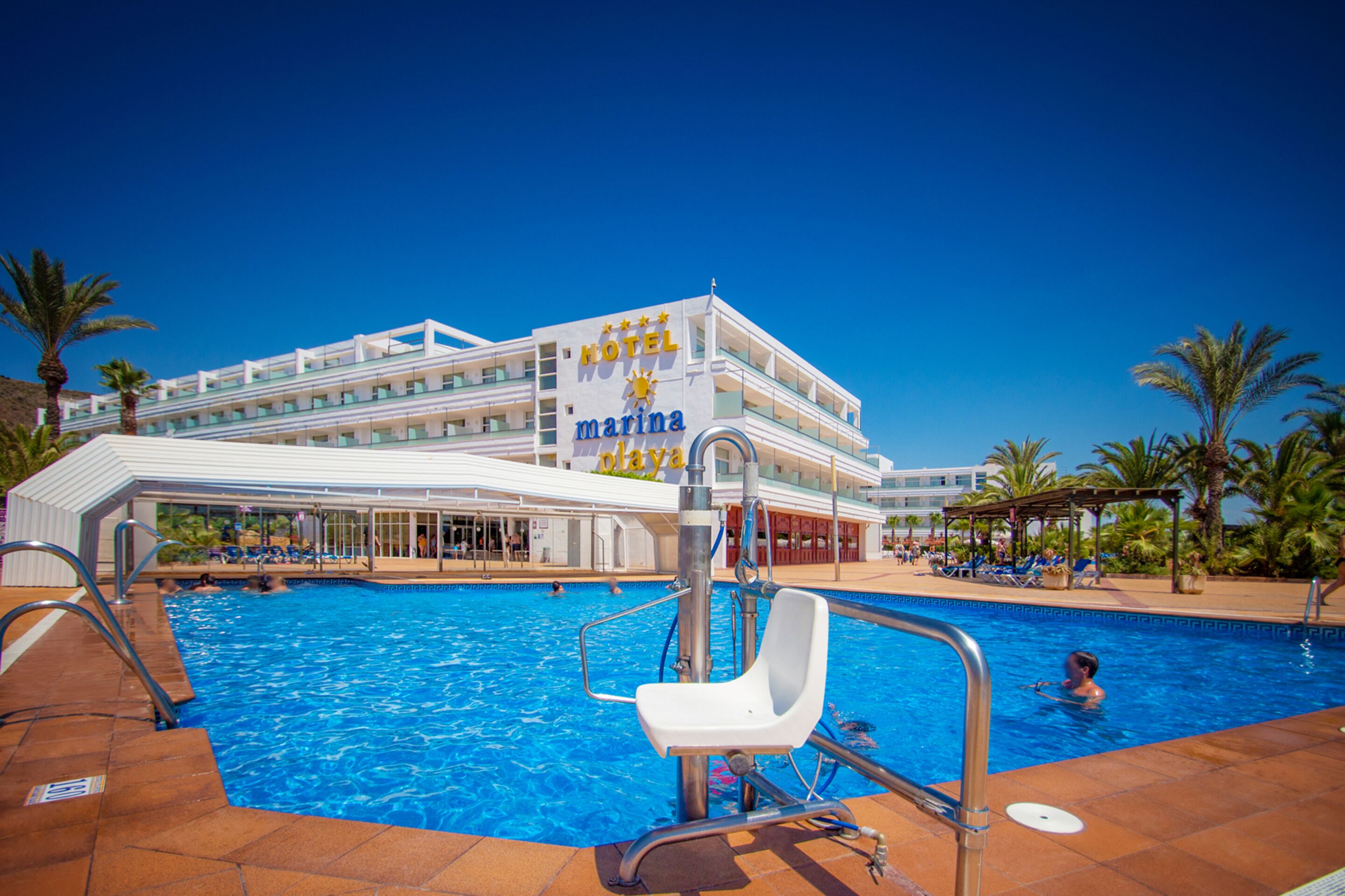 A hotel with a swimming pool, a covered area with lounge chairs, and palm trees.