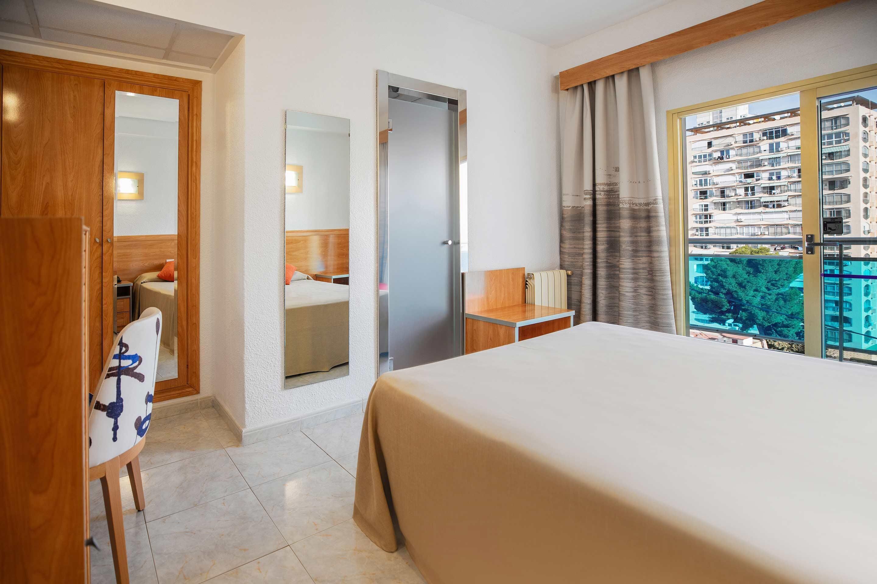 Hotelkamer met een bed, bureau, stoel, kledingkast, spiegel en raam met uitzicht op een gebouw en bomen.