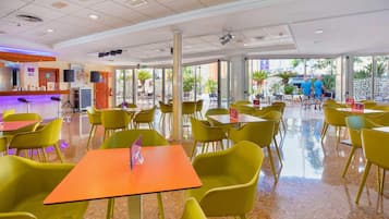 A spacious indoor area with yellow chairs and orange tables, large windows, and a reception desk with a counter and speakers.