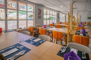 Uma área de refeições moderna com várias mesas e cadeiras, janelas amplas com painéis de vidro decorativos e vista para o exterior.