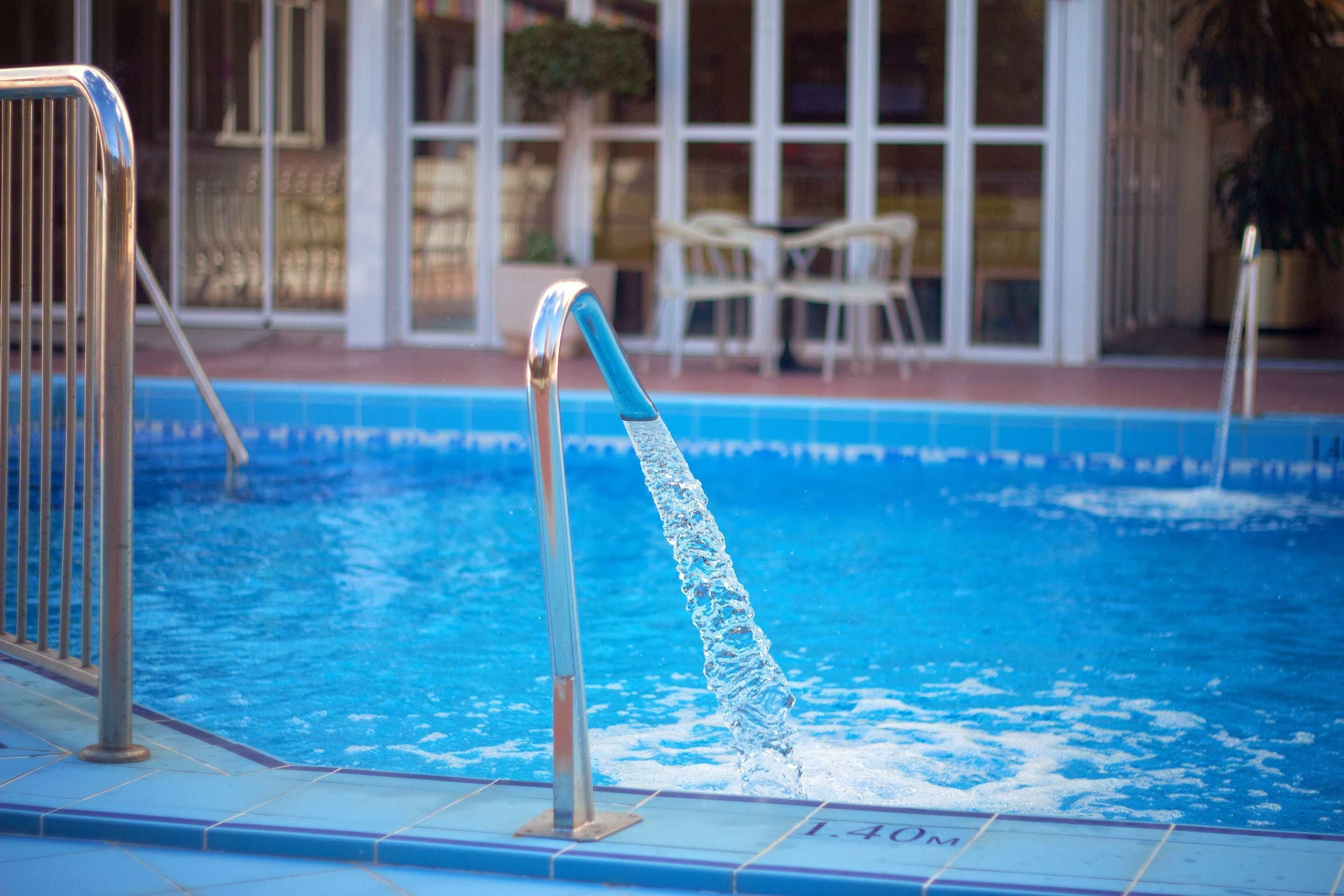 A swimming pool with a water spout, a metal railing, and a tiled deck.