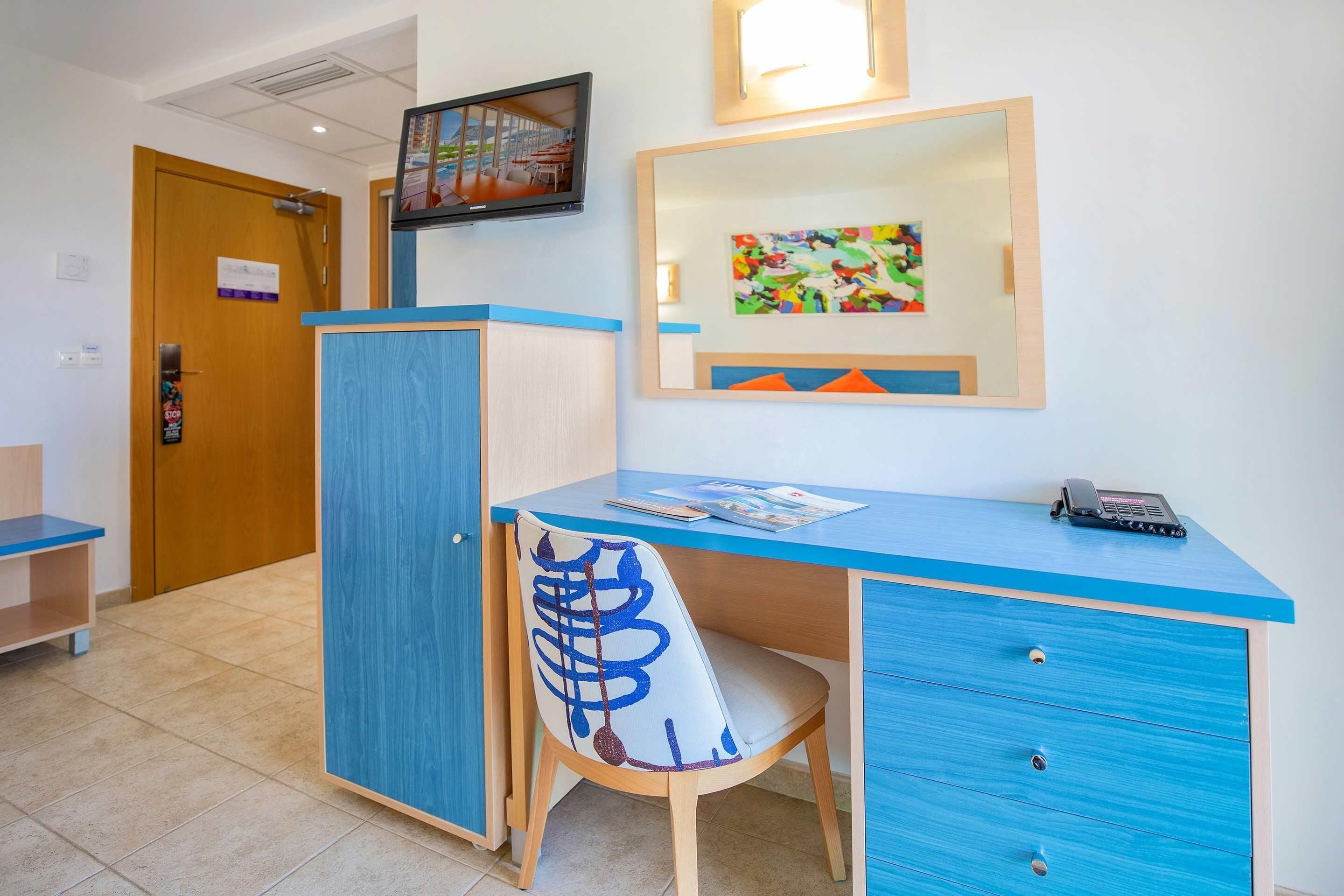 A hotel room with a blue desk, a chair with a unique design, a television mounted on the wall, and a colorful painting above the desk.