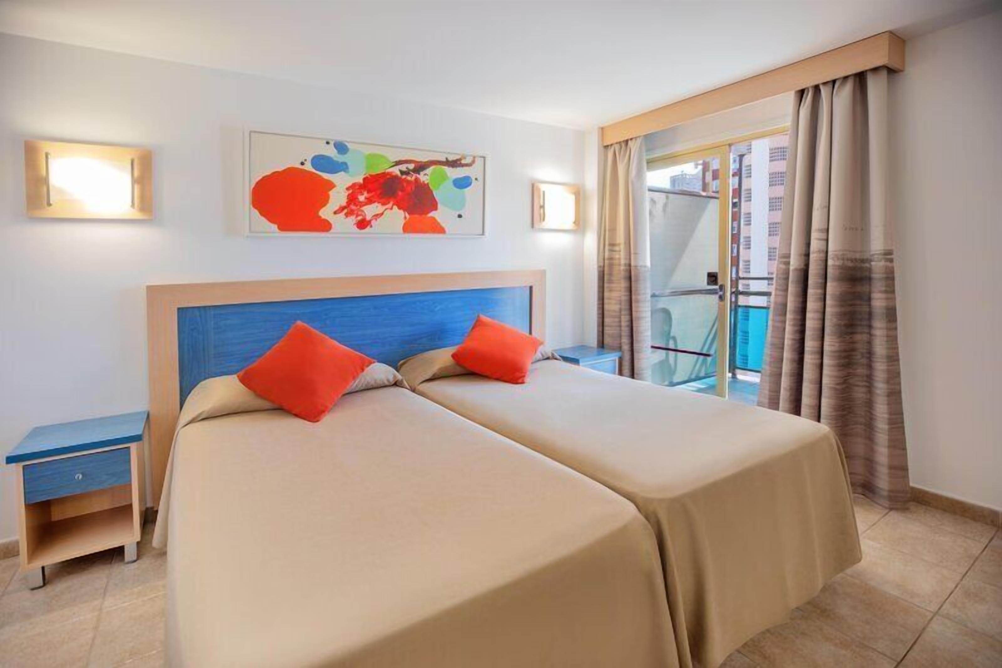 Ein Hotelzimmer mit zwei Betten, einem Nachttisch, einem Sessel und einem Balkon mit Vorhängen, sowie einem Gemälde an der Wand.