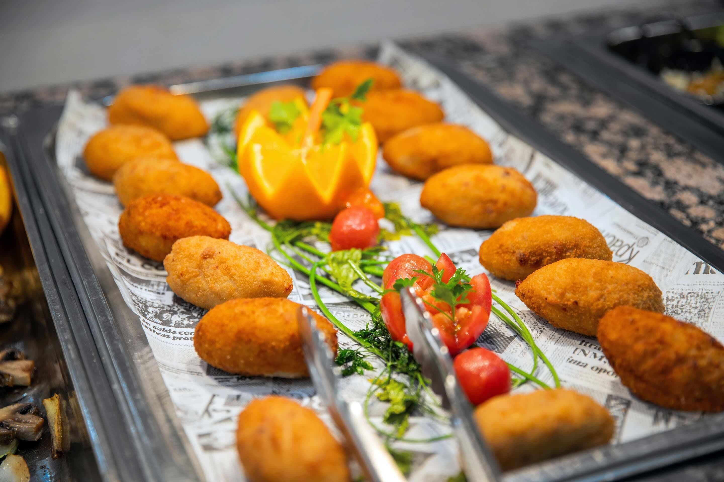 Bandeja con crujientes croquetas de color dorado, dispuestas sobre un papel de periódico, adornadas con tomates cherry, perejil y una rodaja de pimiento.