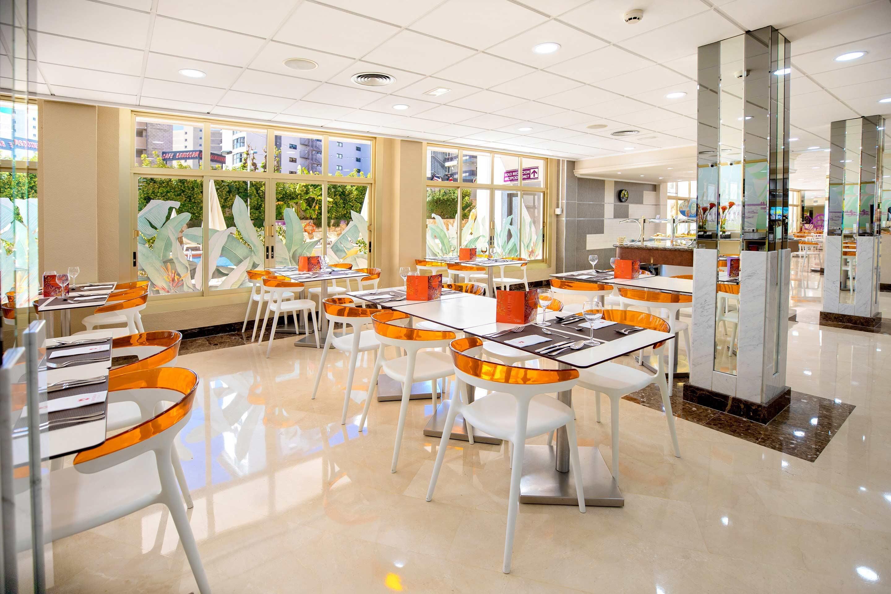 A modern restaurant with glass tables and chairs, large windows with decorative elements, and a clean, polished floor.