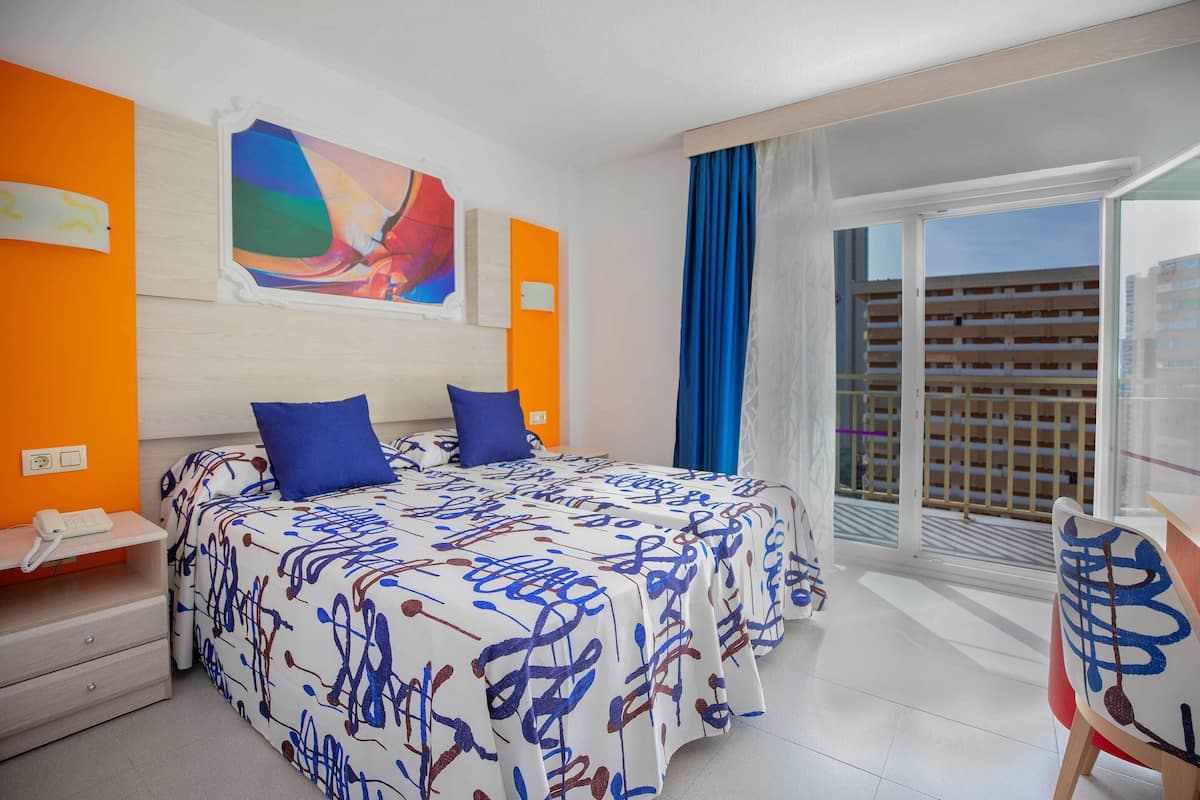 A hotel room with a bed, a desk, and a view of a building through a large window.