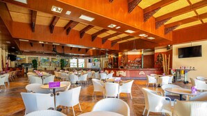 A spacious indoor area with multiple round tables and white wicker chairs, featuring a wooden ceiling and a bar area in the background.
