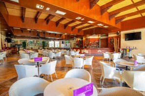 A spacious indoor area with multiple round tables and white wicker chairs, featuring a wooden ceiling and a bar area in the background. - Hotel Servigroup Orange (Benidorm)