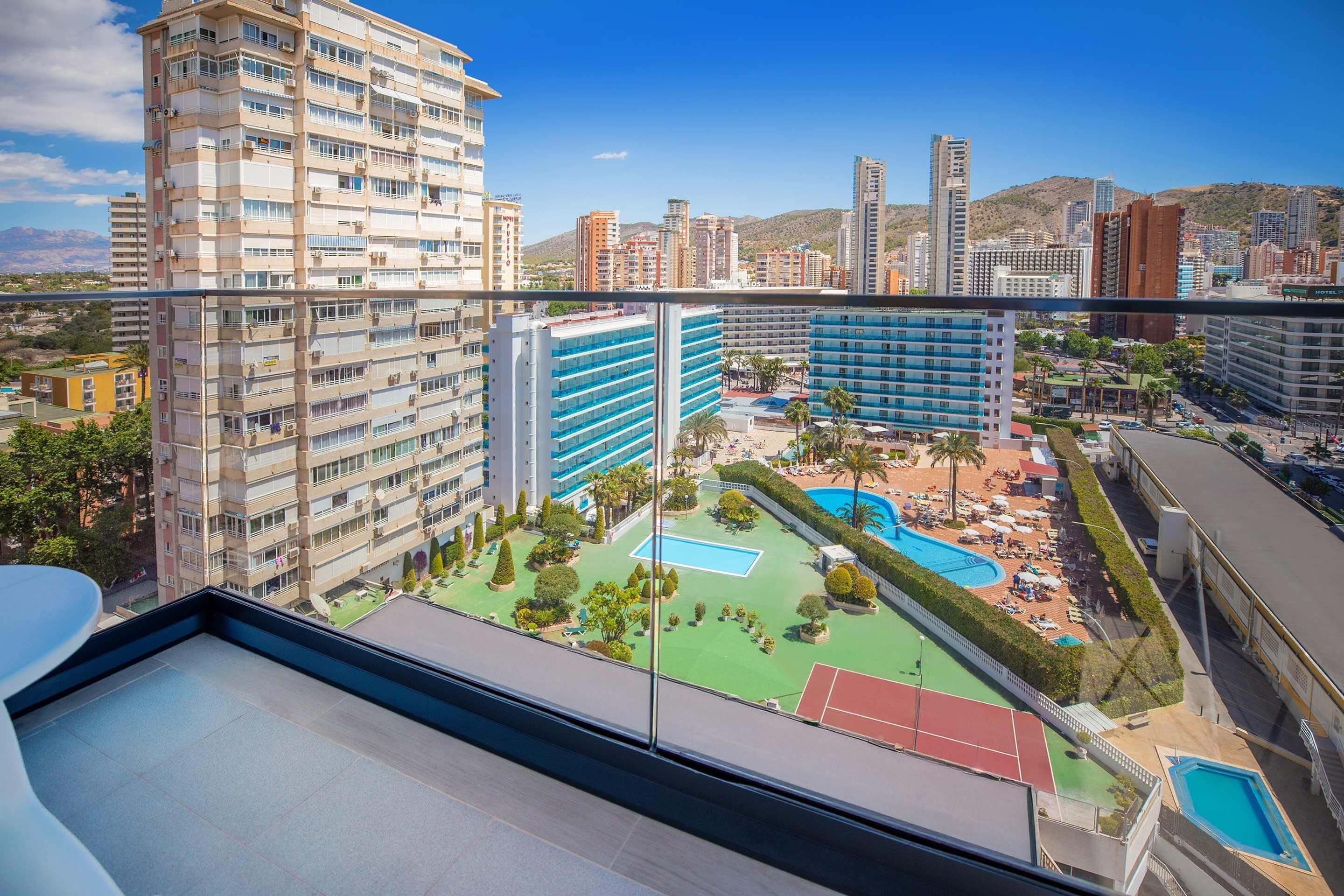 Un hotel alt amb nombroses plantes, una piscina al terrat, pistes de tennis i vista a la ciutat.