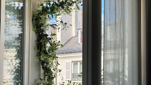 Blick durch ein Fenster mit Vorhang, zeigt einen Balkon mit Geländer und Blumentöpfen.