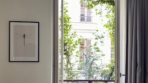 Ein Schlafzimmer mit Bett, einem Sessel, einem Balkon mit Blumentöpfen und einem gerahmten Bild an der Wand.