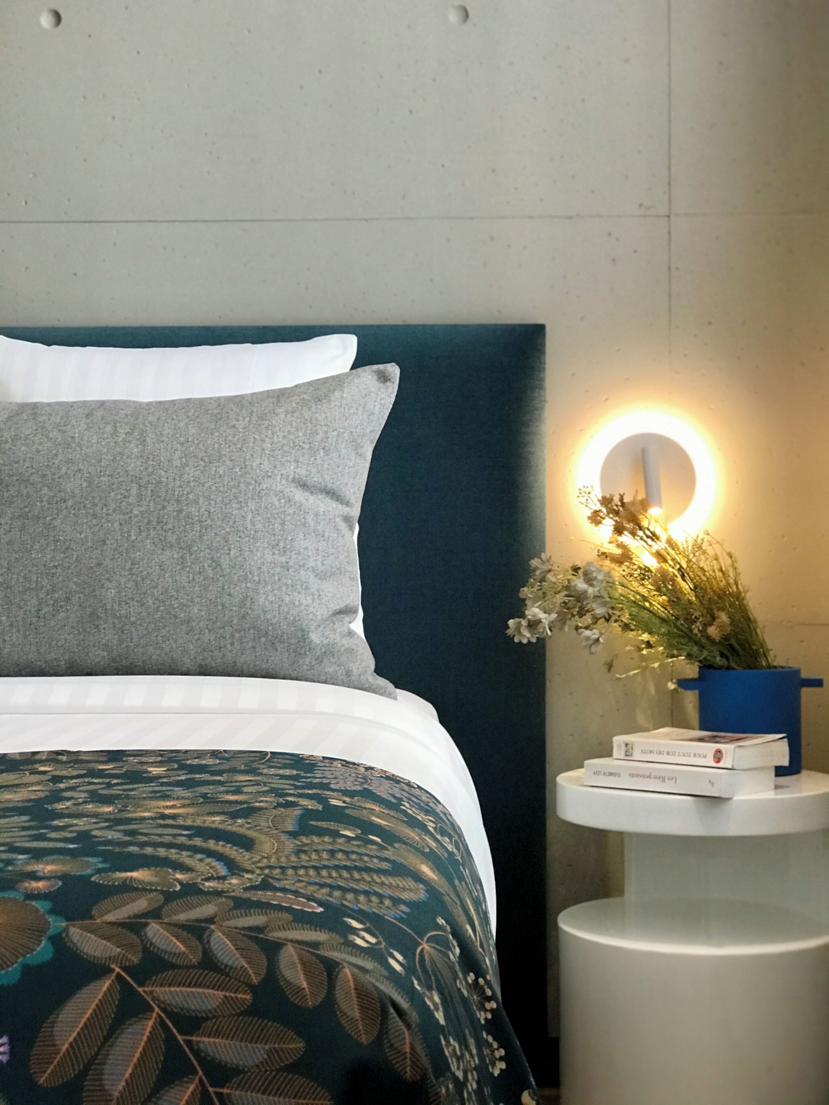 Una cama bien tendida con ropa de cama estampada, una mesita de noche con lámpara y libros, y un aplique en la pared.