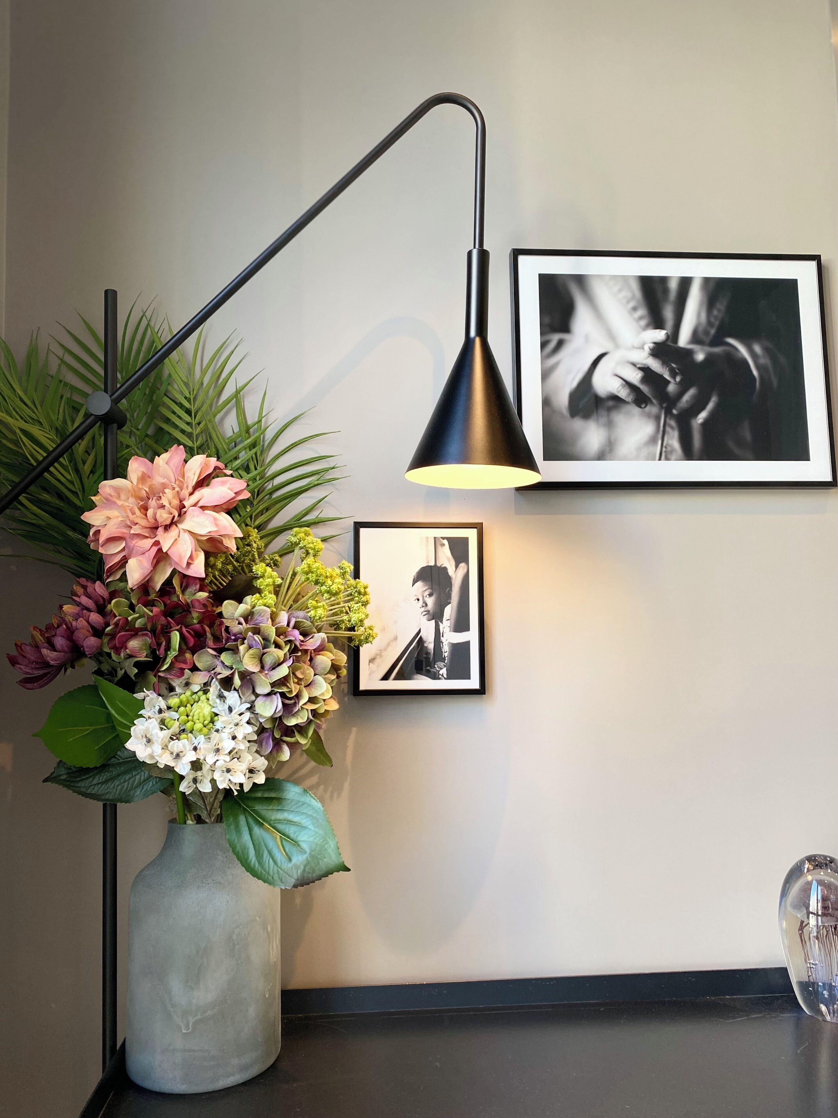 Un escritorio con un florero de flores, una lámpara de escritorio negra y dos fotografías enmarcadas en la pared.