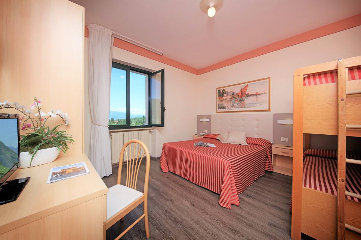 Hotelkamer met een houten bureau, een computer, een stoel, een bed met gestreept beddengoed, een nachtkastje, een bedtafel, een lamp, een raam met gordijnen, een potplant en een schilderij aan de muur.