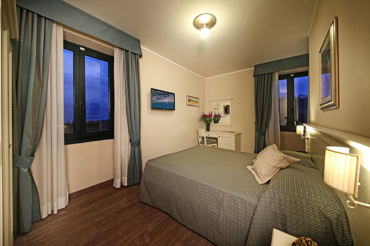 Een hotelkamer met een bed, een bureau, een stoel, een raam met gordijnen, een schilderij en een lamp.