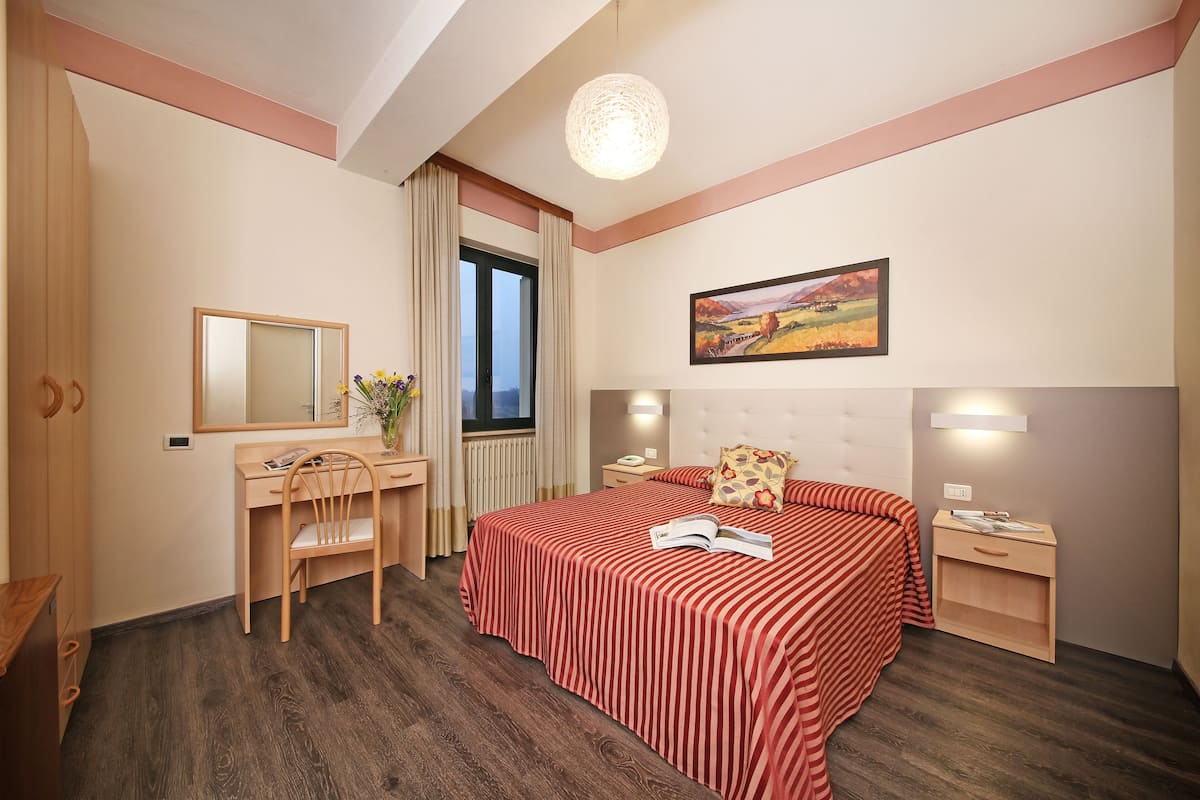 Een slaapkamer met een bed, een bureau, een stoel, een nachtkastje, een bedtafel, een lamp, een raam en een schilderij aan de muur.