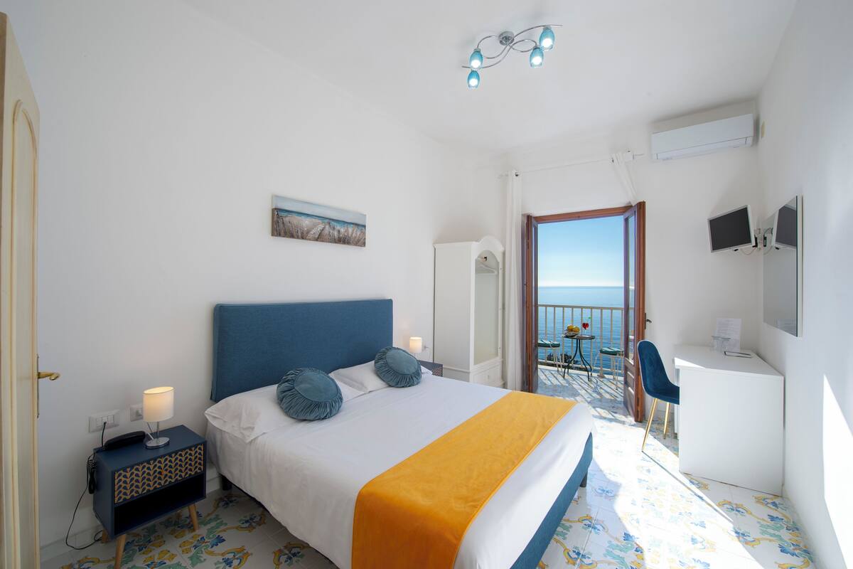 A hotel room with a bed, bedside table, desk, chair, and a balcony with a view of the sea.