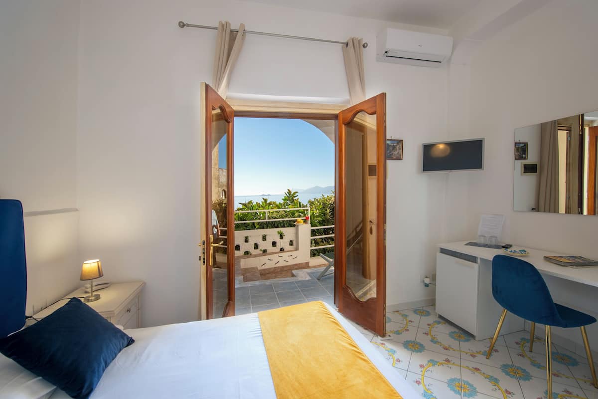 A hotel room with a bed, a desk, a chair, a TV, and a balcony with a view of the sea and mountains.