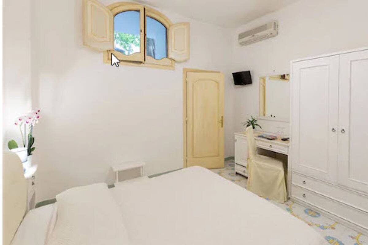 A bedroom with a bed, a wardrobe, a desk, a chair, a small table, a mirror, a window, and a television.