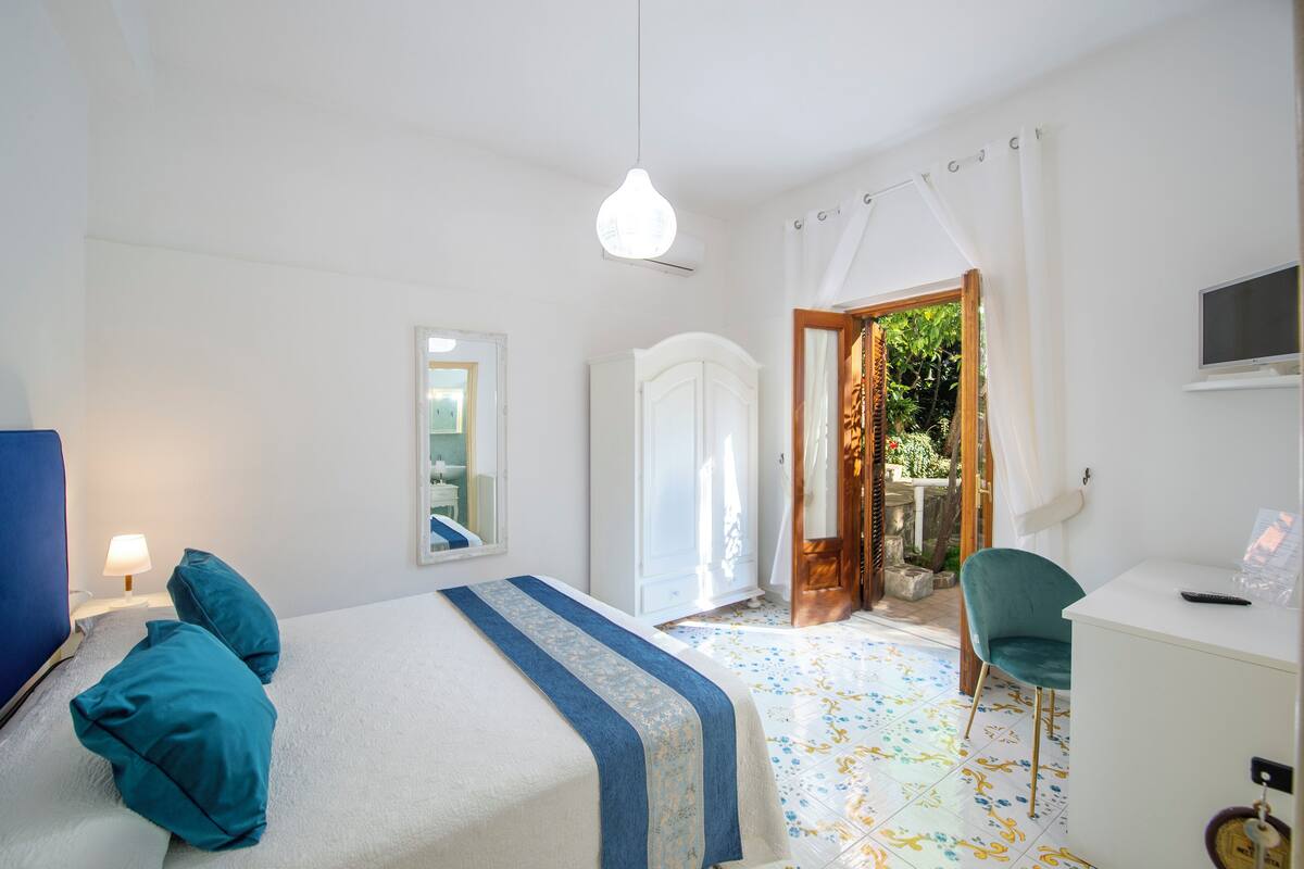 A bedroom with a bed, a desk, a chair, a TV, and a door leading to a garden.