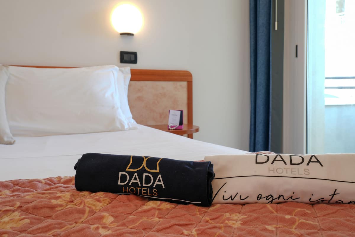 A hotel room with a neatly made bed, a DADA Hotels pillow, a DADA Hotels blanket, and a wall-mounted light.