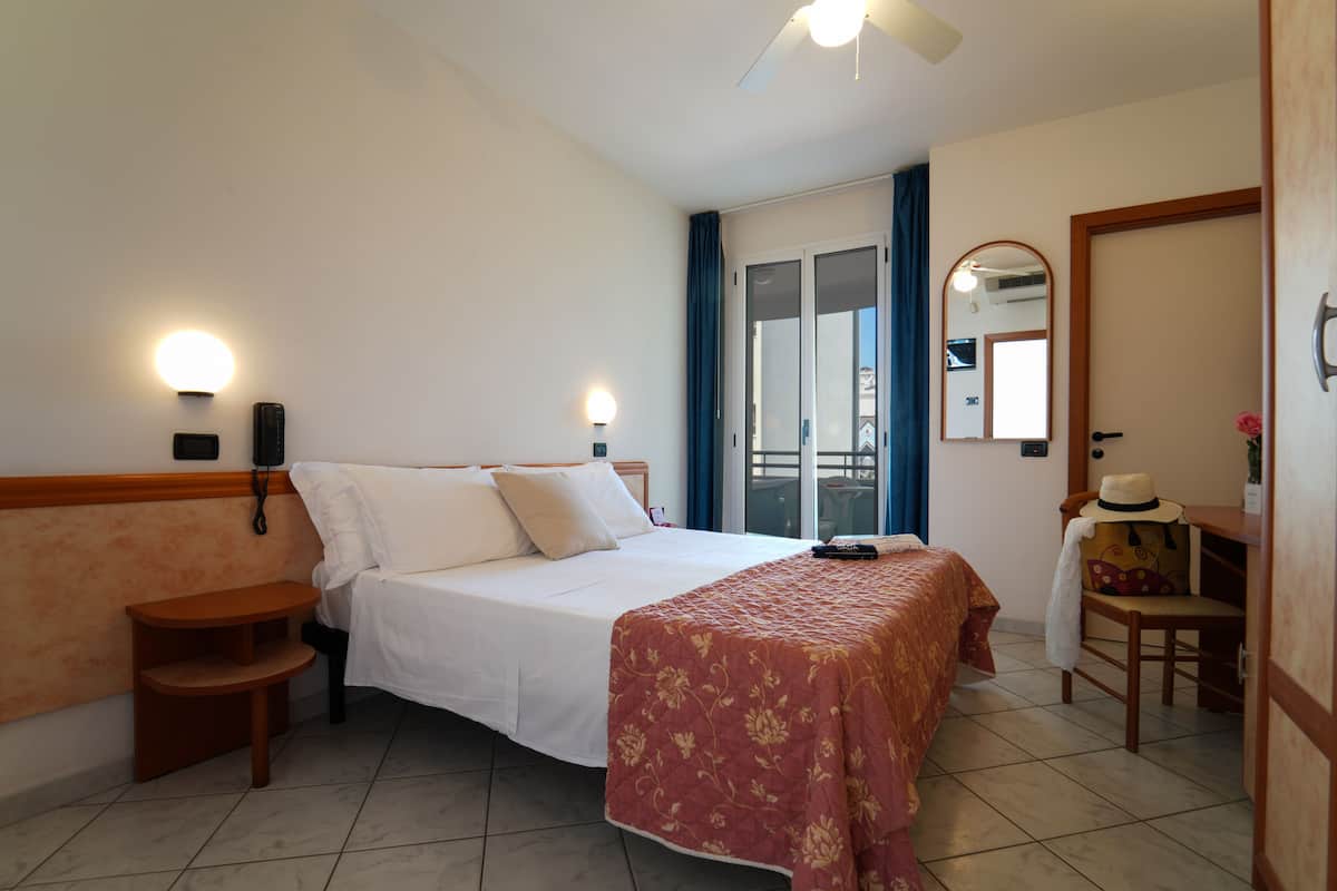 A hotel room with a bed, bedside table, lamp, and a view of the outside balcony.