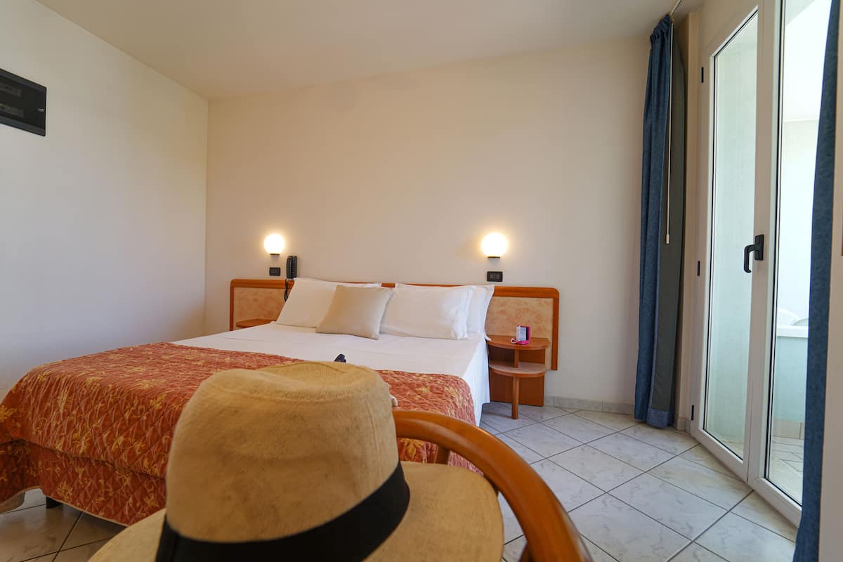 A hotel room with a bed, two bedside tables with lamps, a chair, and a sliding glass door with curtains.