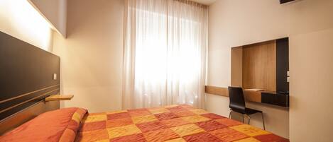 Une chambre d’hôtel dotée d’un grand lit, d’un téléviseur à écran plat fixé au mur, d’un bureau avec une chaise et d’une fenêtre avec des rideaux légers.