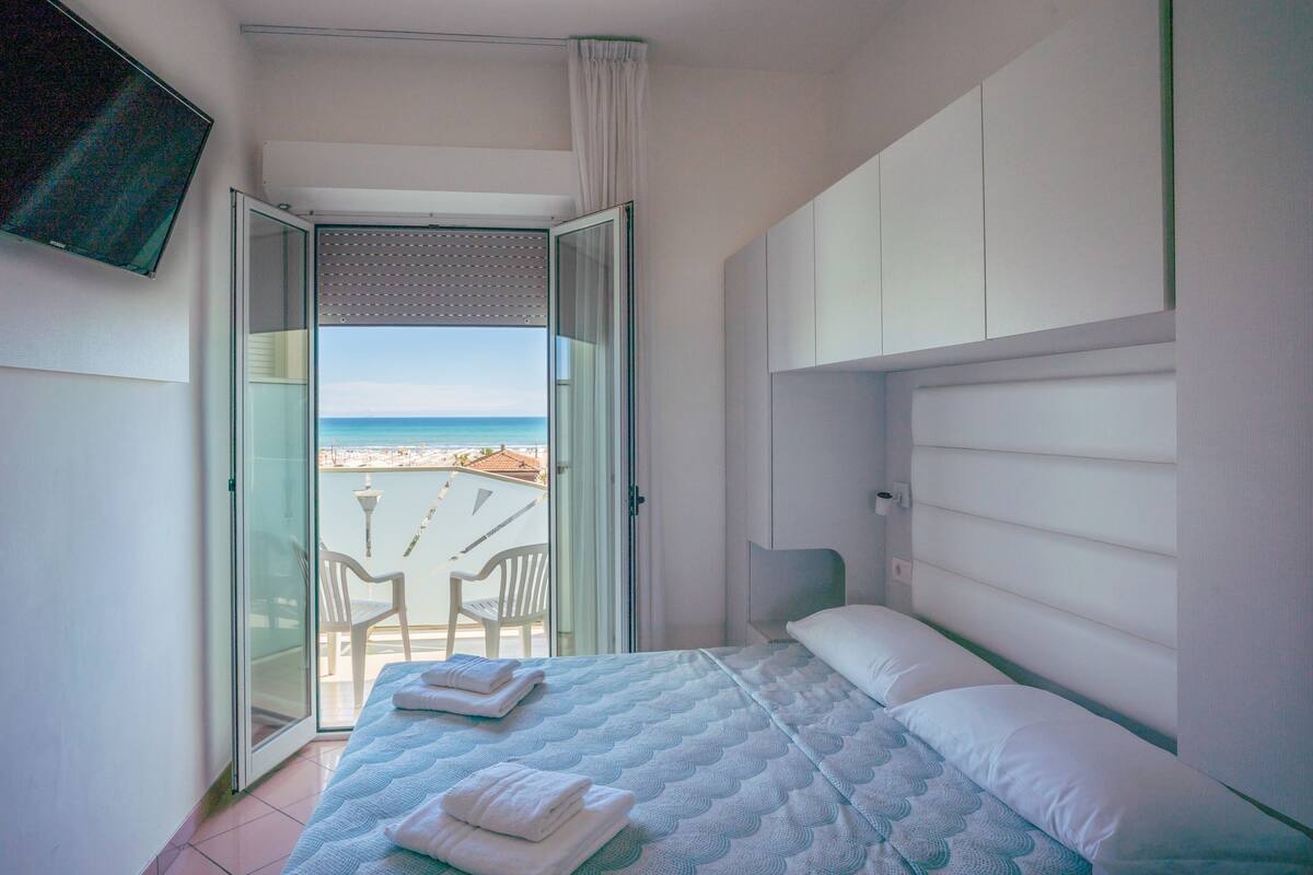 A bedroom with a bed, a TV, and a view of the beach through a sliding door.