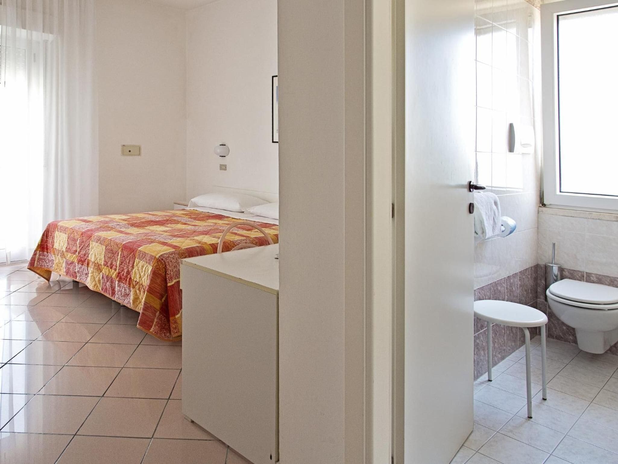 A hotel room with a bed, a small desk, a chair, a toilet, and a window.
