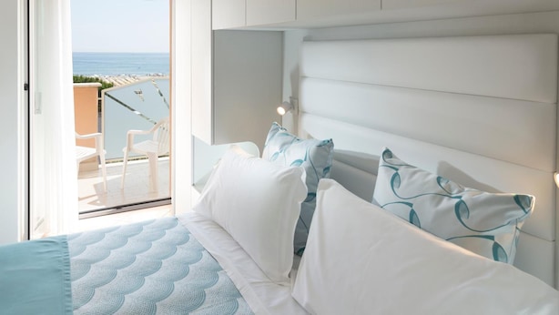 Un dormitorio con una cama grande, cabecera blanca y vistas al mar a través de una puerta abierta.