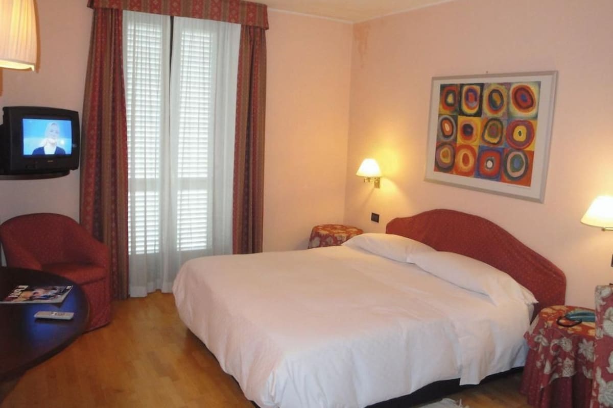 Una camera d'albergo con un letto, due abat-jour, una televisione su un mobile, una sedia e un tavolino con una rivista.