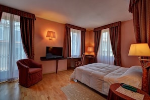 Una camera d'albergo con un letto, una scrivania con televisore, una sedia e un piccolo tavolo con lampada.