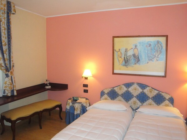 A hotel room with a bed, a bench, a small table, a lamp, and a painting on the wall.