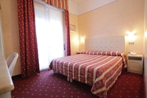 Una habitación de hotel con una cama, una mesita de noche, una silla y un ventanal con cortinas.
