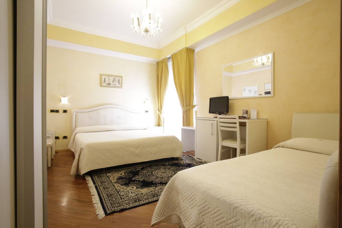 A hotel room with two beds, a desk with a computer, a chandelier, and a large mirror.