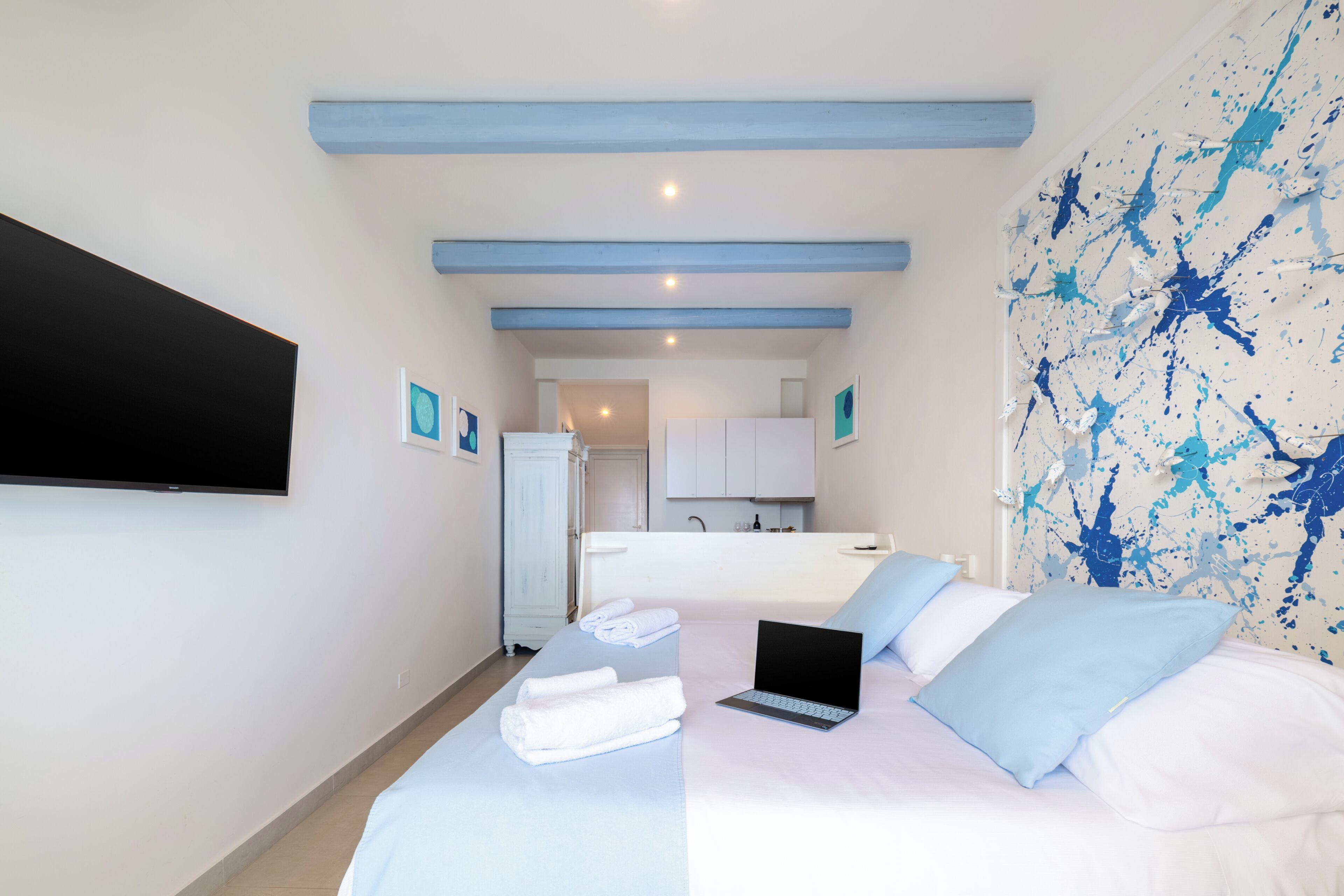 A bedroom with a bed, a laptop, towels, a TV, and a bathroom visible in the background.