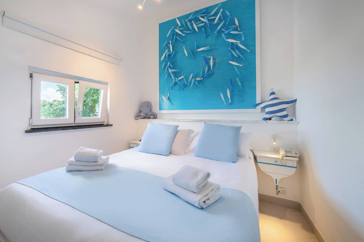 A bedroom with a bed, pillows, a window, a bedside table, and a wall art piece.