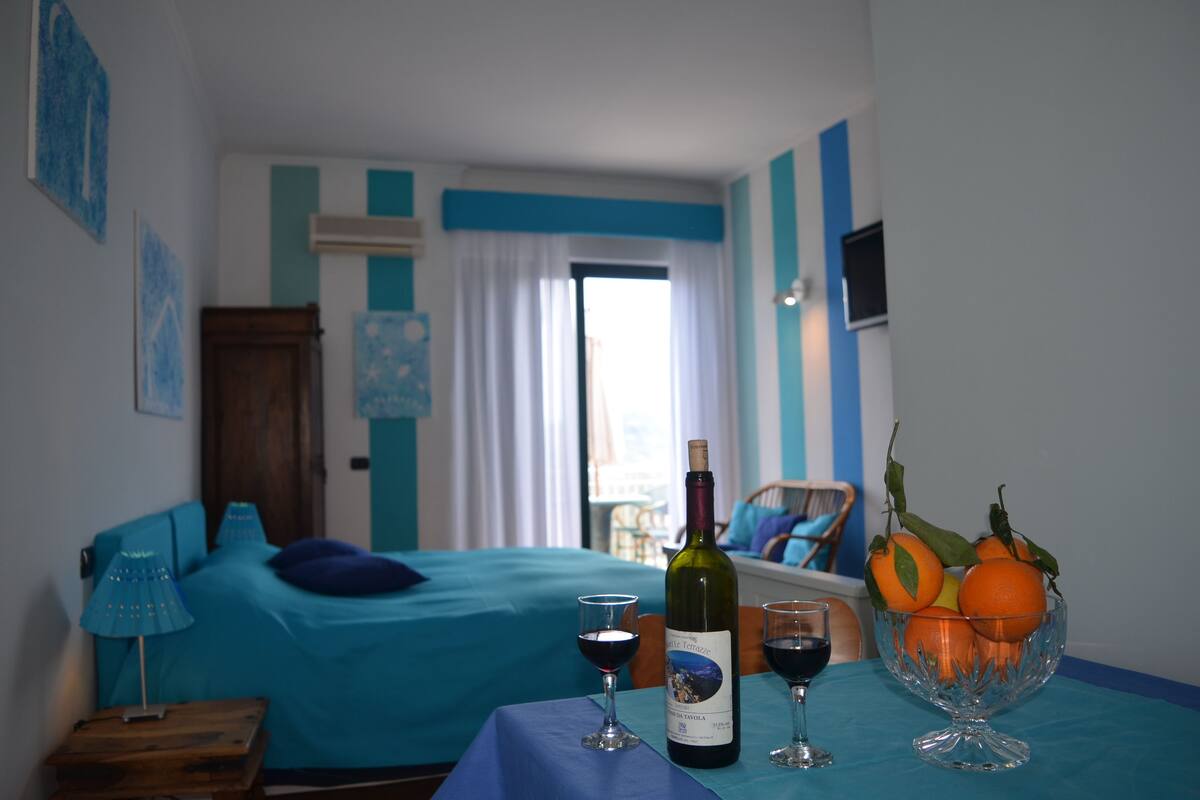 A hotel room with a bed, a table with a bottle of wine, glasses, and a bowl of fruit.