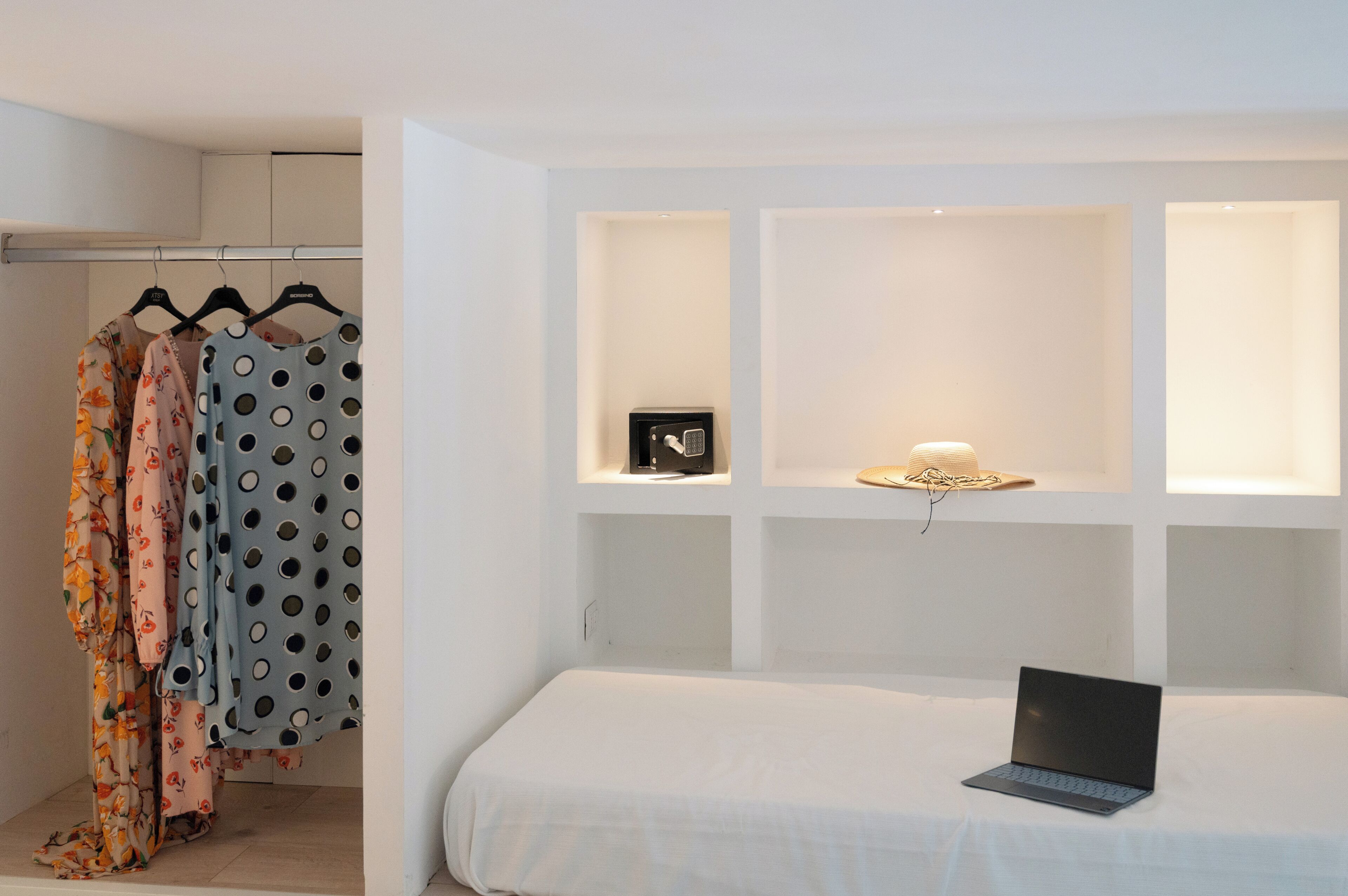 A bedroom with a bed, a laptop, a wardrobe with clothes, and a shelf with a lamp and a safe.