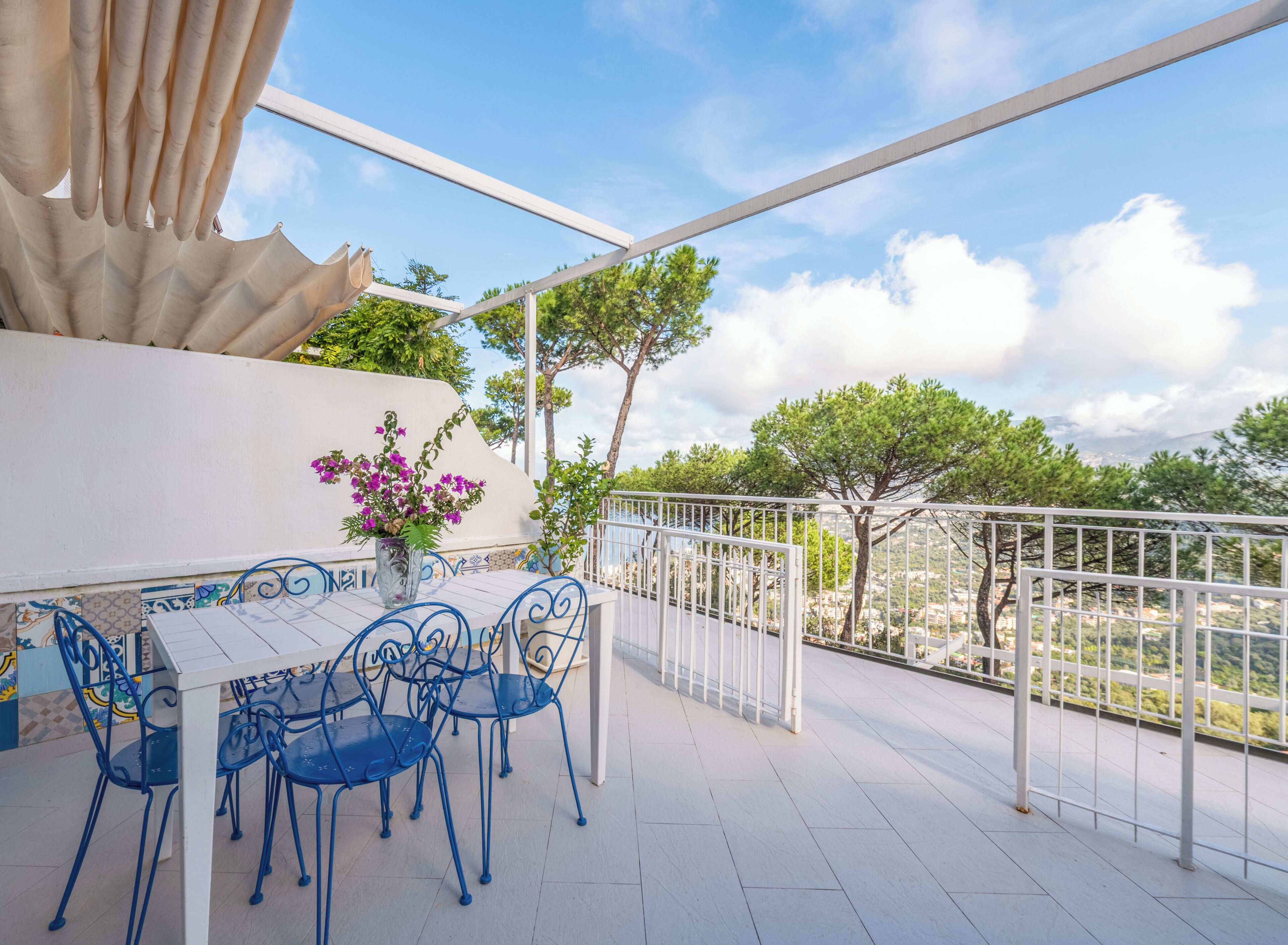 An outdoor terrace with a table and chairs, a white railing, and a view of trees and buildings.