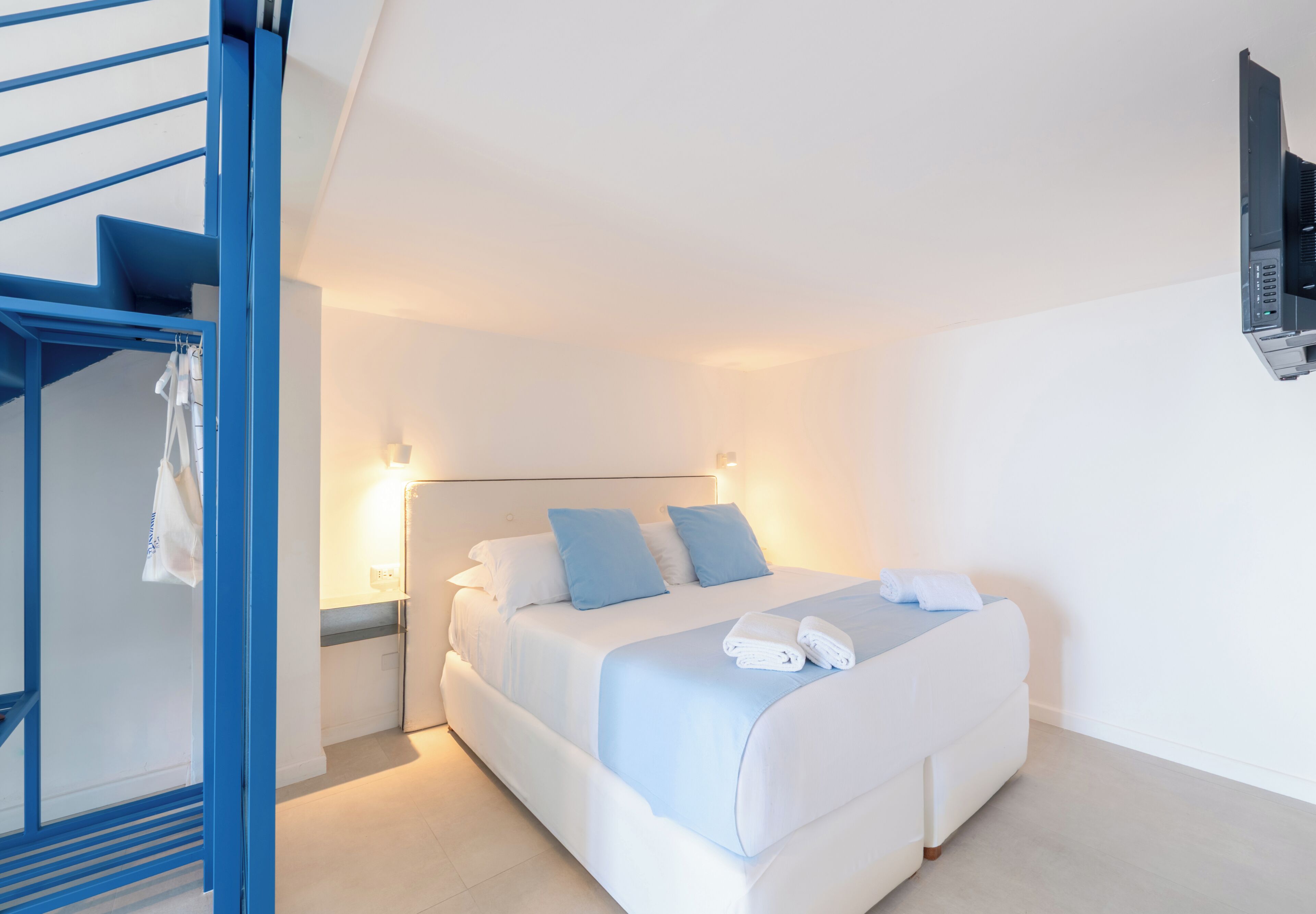 Quarto com cama, escadaria azul e TV fixada na parede.