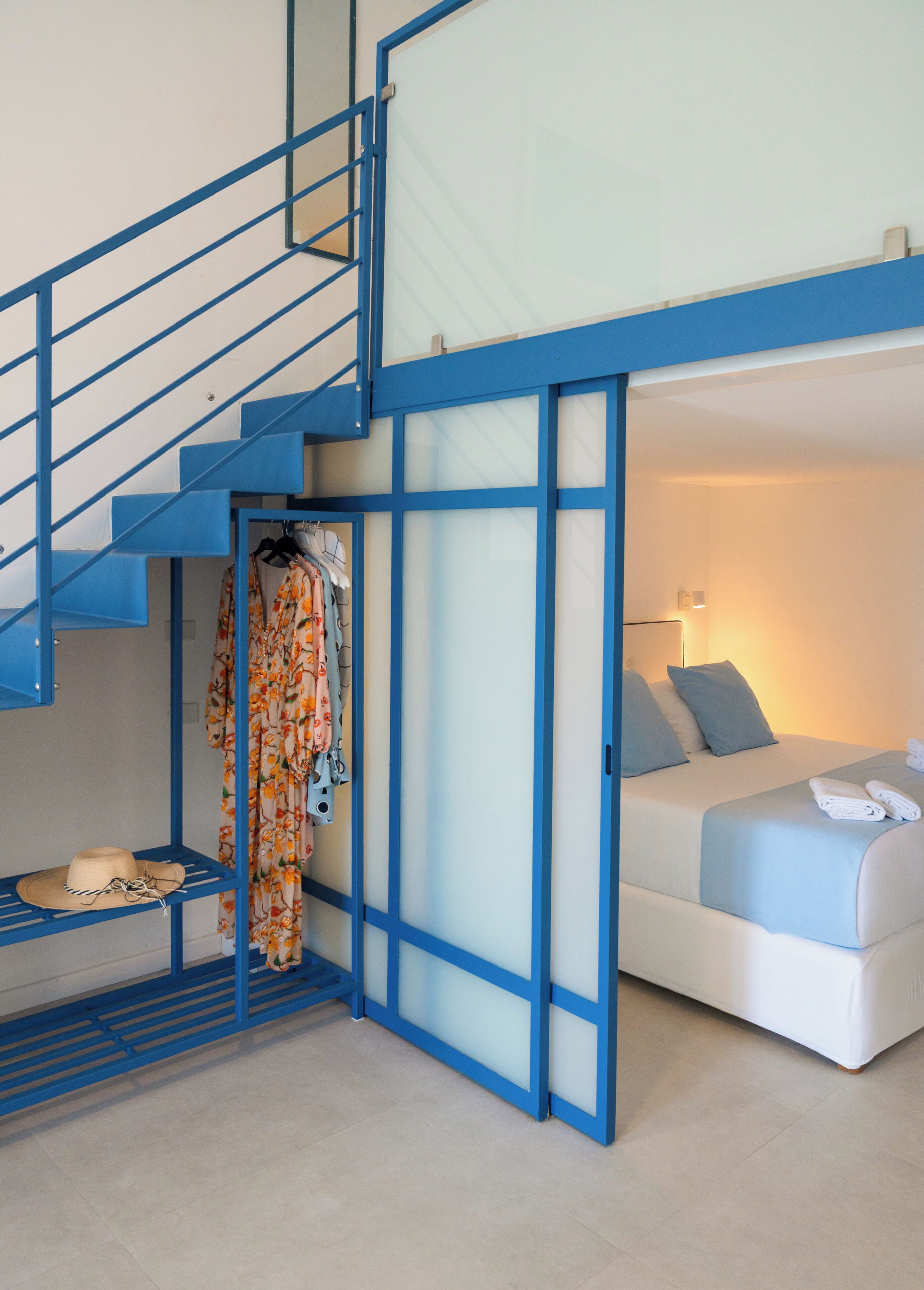 A modern hotel room with a blue staircase, a bed with pillows, and a wardrobe with hanging clothes.