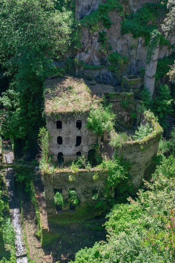 A historic stone building with multiple arched openings, surrounded by lush greenery and perched on a cliffside.