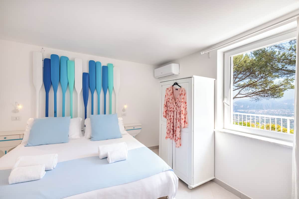 A bedroom with a bed, pillows, a wardrobe, a window with a view, and a wall-mounted air conditioner.