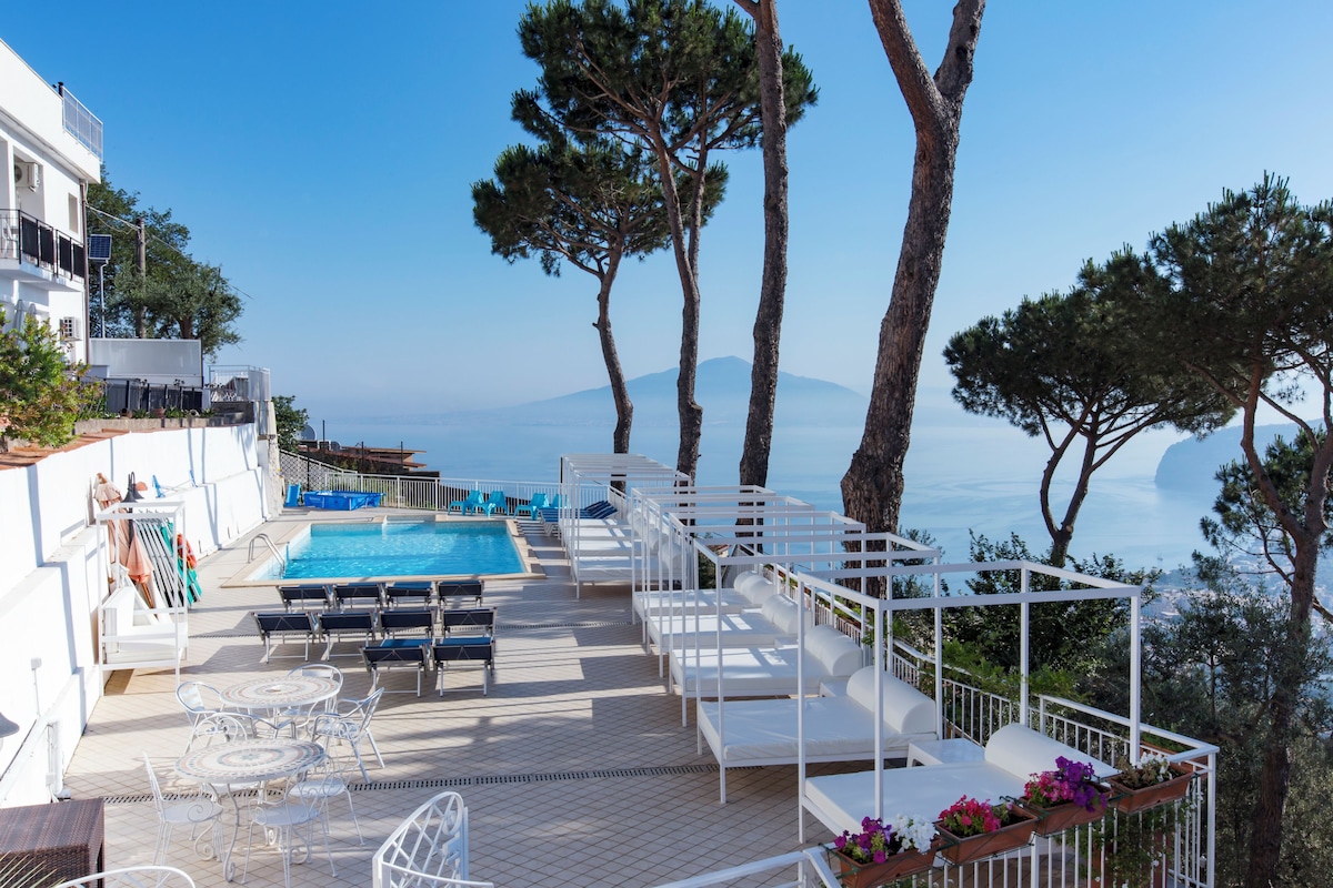 A rooftop pool area with lounge chairs and tables, overlooking a scenic view of the sea and mountains.
