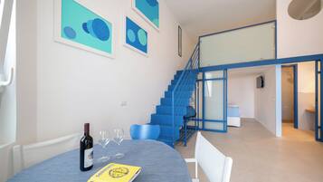 Un área de comedor con una mesa dispuesta para dos, una botella de vino y un menú. Una escalera con barandas azules conduce a un nivel superior.