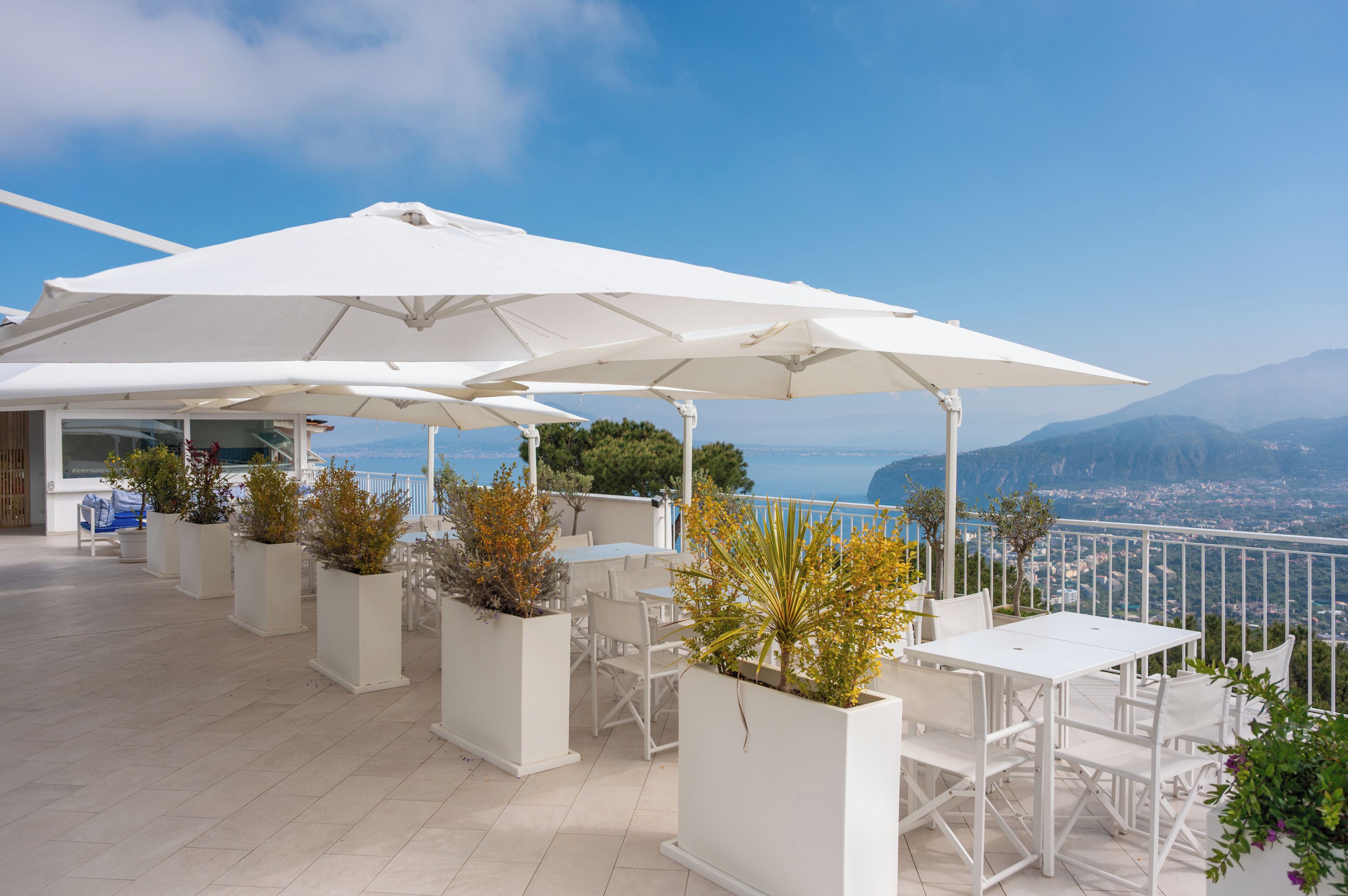 Una terraza con mobiliario blanco, sombrillas y plantas en macetas.