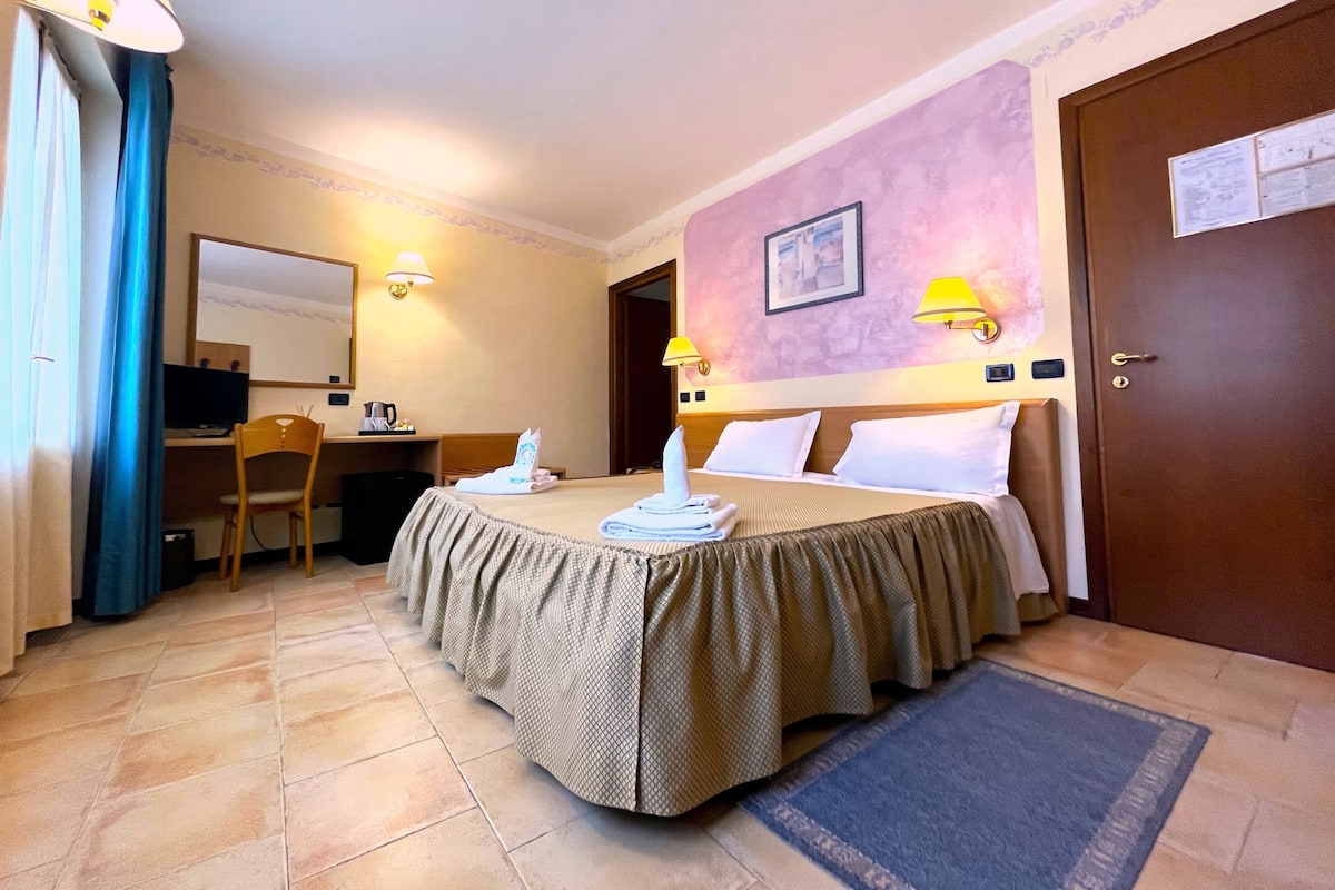 A hotel room with a bed, a desk with a computer, a chair, and a desk lamp.